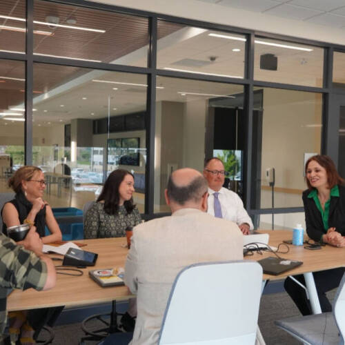 A group of six professionals sit around a conference table engaged in a discussion inside a modern office space with large windows and glass walls. The group includes both men and women, and they appear to be collaborating in a meeting.